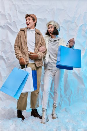 amazed interracial couple in winter attire with shopping bags looking away on white snow in studio