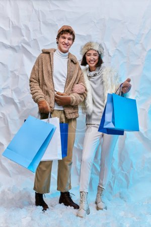 zufriedenes gemischtrassiges Paar in warmer Kleidung mit Einkaufstüten im verschneiten Studio vor der Kamera