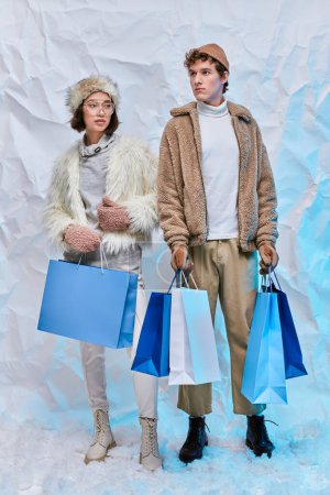 Wintereinkauf, multiethnisches Paar in saisonaler Kleidung mit weißen Einkaufstaschen auf Schnee im Atelier