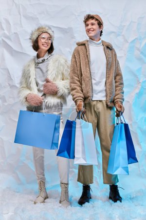 campaña de moda de invierno, feliz pareja interracial con bolsas de compras azules en la nieve blanca en el estudio