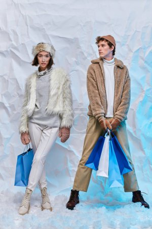 trendy interracial couple posing with blue shopping bags on white snow in studio, winter fashion