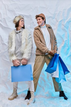 trendy interracial couple with shopping bags smiling at each other on snow in studio, winter style