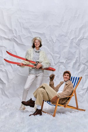 cheerful asian woman with skis near trendy man in deck chair on snow in studio, winter leisure