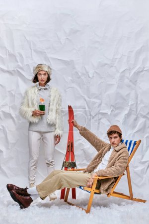 stylischer Mann mit Skiern sitzt im Liegestuhl neben asiatischer Frau mit heißem Cocktail auf Schnee im Studio