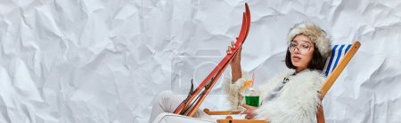 Photo for Asian woman in winter attire in deck chair with skis and cocktail on white textured backdrop, banner - Royalty Free Image
