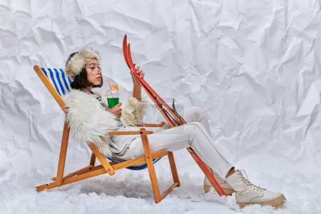Photo for Asian woman in warm trendy outfit with skis and cocktail relaxing in deck chair on snow in studio - Royalty Free Image