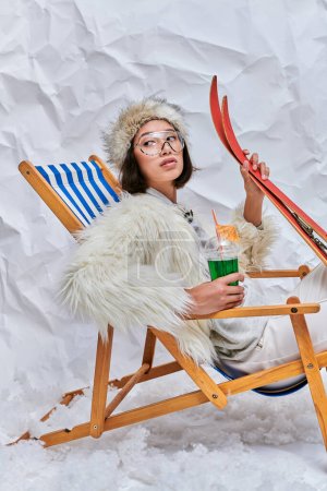 Asiatin mit Brille und Winterbekleidung entspannt im Liegestuhl mit Cocktail und Skier im Studio