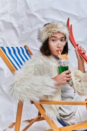 Photo for Asian woman with skis drinking hot teddy cocktail in deck chair on white textured backdrop - Royalty Free Image