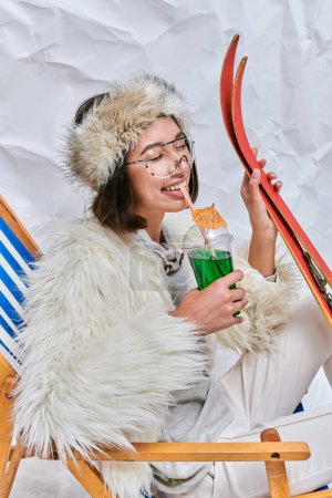Photo for Happy asian woman with skis drinking hot teddy cocktail with closed eyes on white textured backdrop - Royalty Free Image