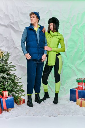 multiethnisches Paar in warmer Kleidung in der Nähe von Geschenkboxen und Weihnachtsbaum auf Schnee im Atelier, Weihnachtszeit