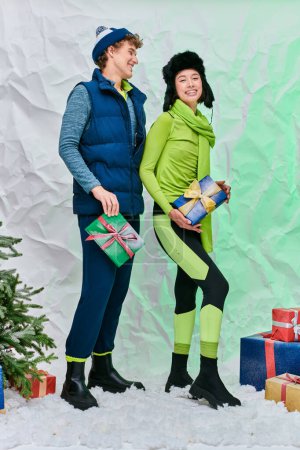 freudiges gemischtrassiges Paar in hellen Kleidern posiert mit Weihnachtsgeschenken im festlich verschneiten Studio
