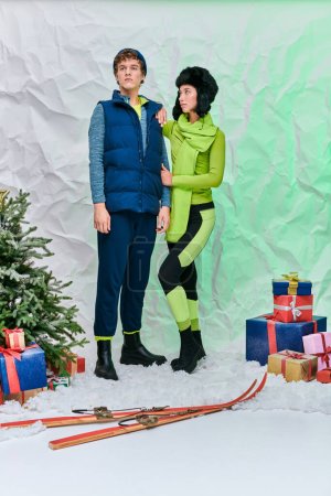 Modernes Paar in Winterbekleidung in der Nähe von Geschenken, Skiern und Weihnachtsbaum auf Schnee im Atelier