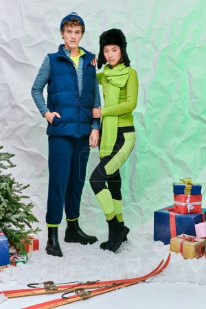 pareja interracial de moda en ropa de abrigo cerca de regalos, esquís y árbol de Navidad en el estudio nevado