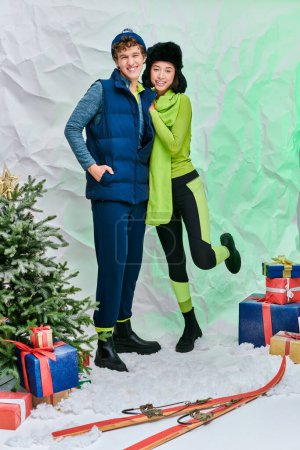 interracial couple in winter outfit smiling near christmas tree and presents on snow in studio