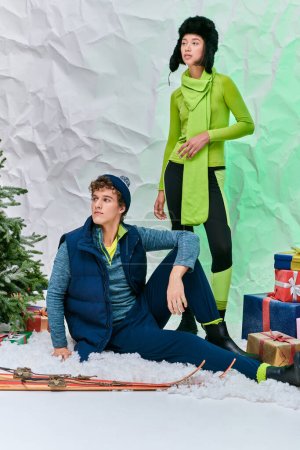 interracial couple in winter wear looking away next to presents and christmas tree in snowy studio