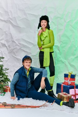 Paar in Winterkleidung lächelt neben Weihnachtsbaum, Skiern und Geschenken im verschneiten Atelier