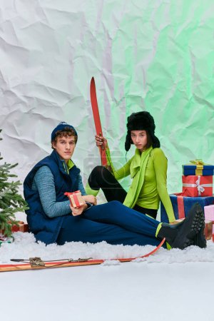 junge asiatische Frau mit Skiern sitzt neben trendigem Mann, Weihnachtsbaum und Geschenken im verschneiten Studio