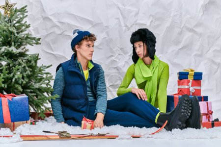 Modisches Paar sitzt auf Schnee im Atelier neben Skiern, Geschenken und Weihnachtsbaum