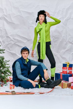 Téléchargez les photos : Vibrant couple interracial regardant caméra près de cadeaux et arbre de Noël en studio enneigé - en image libre de droit