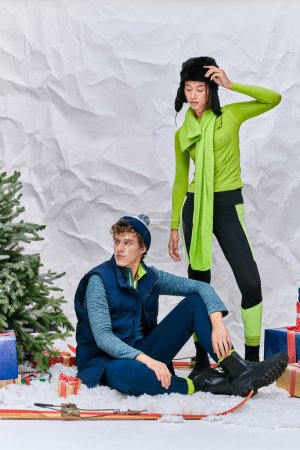 trendiger Mann sitzt auf Schnee im Atelier neben asiatischer Frau, Geschenkschachteln und Weihnachtsbaum, Wintermode