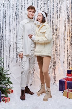 feliz asiático mujer mirando con estilo hombre cerca de regalos y navidad árbol en brillante tinsel telón de fondo