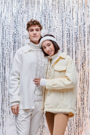 joyful interracial couple in white jackets standing near silver tinsel on backdrop, christmas spirit
