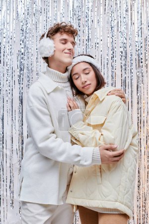 glückliches gemischtrassiges Paar in gemütlicher Winterkleidung, das sich mit geschlossenen Augen in der Nähe von glänzendem Lametta im Studio umarmt