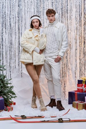 trendy interracial models looking at camera near christmas tree and presents on shiny backdrop