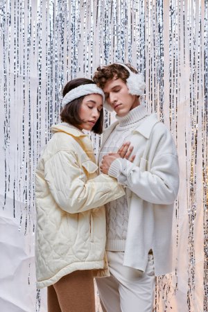espíritu navideño, pareja interracial de moda abrazando con los ojos cerrados sobre el telón de fondo de oropel brillante