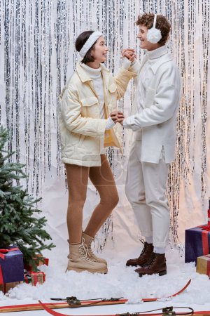 glückliches gemischtes Paar, das sich in der Nähe von Weihnachtsbaum, Geschenkschachteln und Lametta im Atelier anschaut