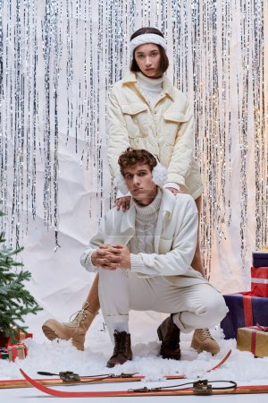 Foto de Pareja multiétnica en ropa acogedora mirando a la cámara cerca del árbol de Navidad, regalos y oropel de plata - Imagen libre de derechos