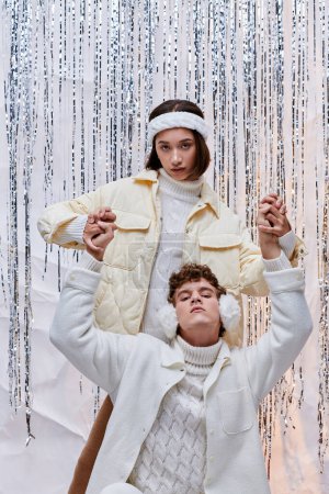 pareja interracial de moda tomados de la mano mientras posan cerca de oropel brillante en el estudio, estilo de invierno