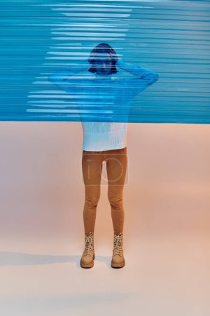 Photo for Full length of young asian woman in white sweater and leather boots behind blue plastic - Royalty Free Image