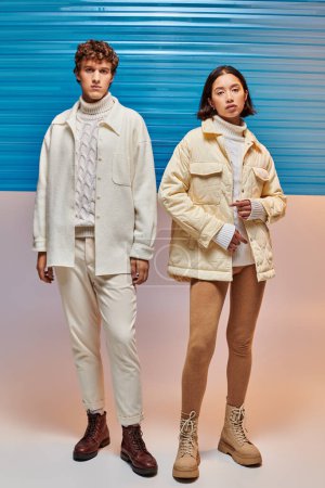 full length of interracial couple in warm jackets and leather boots posing near blue plastic sheet