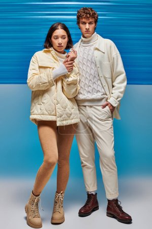 stylischer Mann mit Hand in Tasche neben asiatischer Frau in Winterbekleidung auf blauem Hintergrund mit Plastikfolie