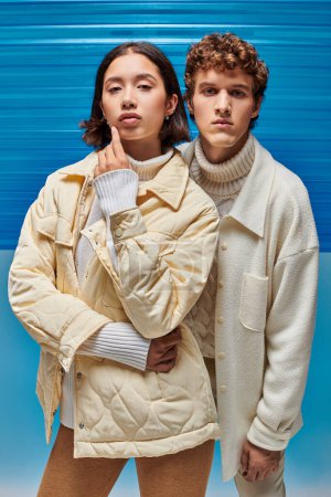 campagne de mode d'hiver, vestes de couple interracial regardant la caméra sur fond de plastique bleu