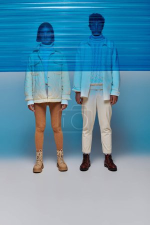 Foto de Pareja interracial en chaquetas y botas de cuero de pie detrás del panel de plástico azul, estilo invierno - Imagen libre de derechos