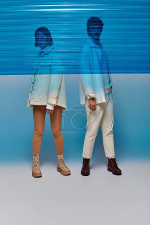 multiethnisches Paar in Jacken und Lederstiefeln posiert hinter blauem Plastiktuch, Wintermode