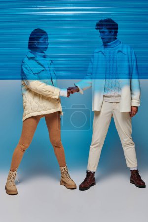 full length of stylish interracial couple holding hands behind blue plastic in studio, winter outfit
