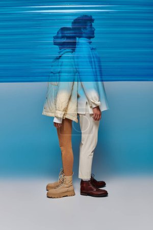 Foto de Vista lateral de pareja interracial en ropa de abrigo de pie espalda con espalda detrás de plástico azul en el estudio - Imagen libre de derechos