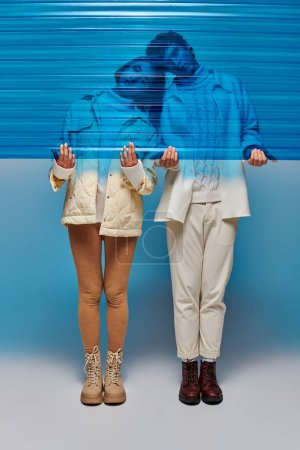 pareja interracial en botas de cuero sosteniendo lámina de plástico azul en el estudio, concepto de moda de invierno