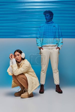 Photo for Asian woman in earmuffs sitting on haunches near man behind blue plastic sheet, winter fashion - Royalty Free Image