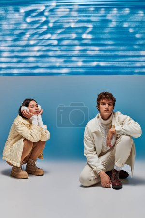 pareja interracial vestida a la moda posando en haunches cerca de la lámina de plástico azul en el estudio