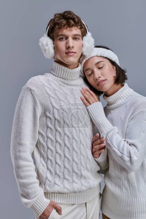 glückliche asiatische Frau in weißem Pullover und Stirnband lehnt auf der Brust des Mannes in warmen Ohrenschützern auf grau