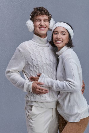 fröhliches multiethnisches Paar in Strickpullovern, das sich bei Schneefall vor grauer Kulisse im winterlichen Stil umarmt