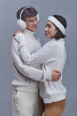 glückliches multiethnisches Paar in warmen Pullovern, das sich unter fallenden Schneeflocken auf grauem Hintergrund umarmt