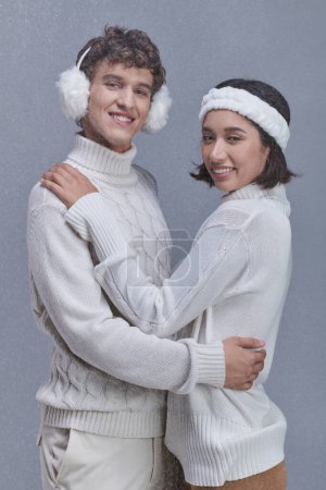 trendy interracial couple embracing and smiling camera on grey background with snow, winter style