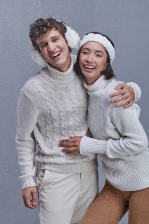 aufgeregtes gemischtes Paar in warmen Pullovern umarmt und lacht unter fallendem Schnee, froher Winter
