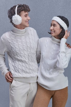 zufriedenes multiethnisches Paar in weißen Pullovern, die einander auf grauem Hintergrund mit Schnee anschauen