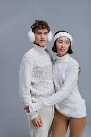 homme en cache-oreilles regardant caméra près de jeune femme asiatique sur fond gris avec de la neige, hiver romantique